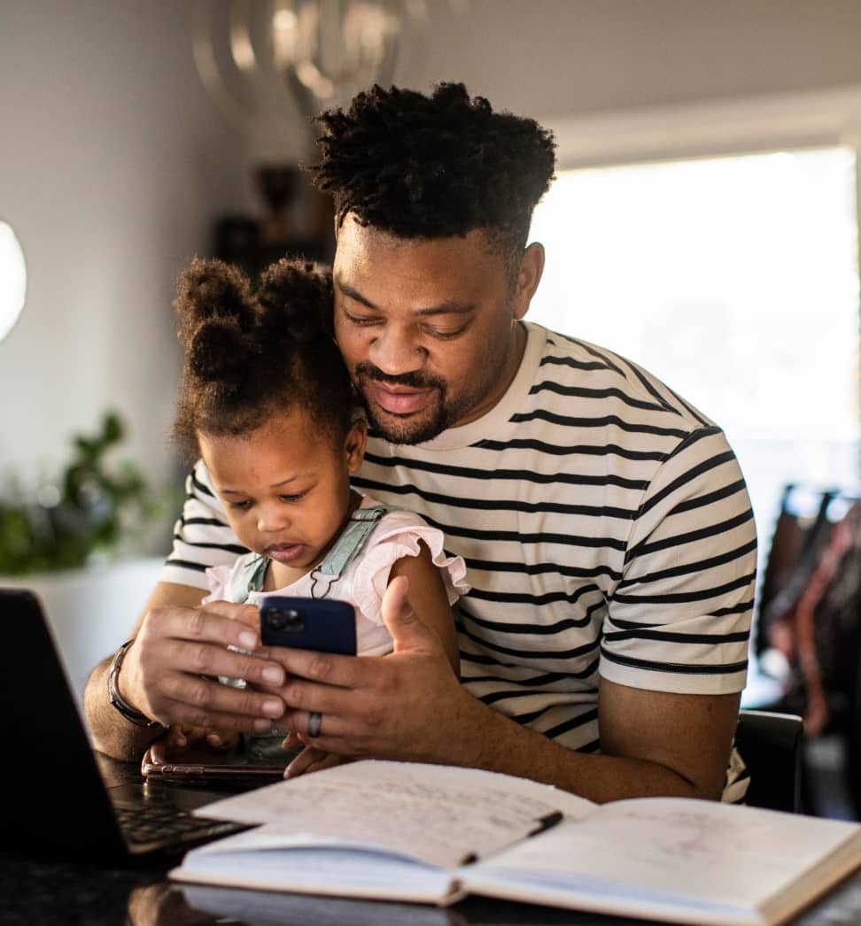 Man on his phone working with his laptop on the counter, with his daughter sitting on his lap